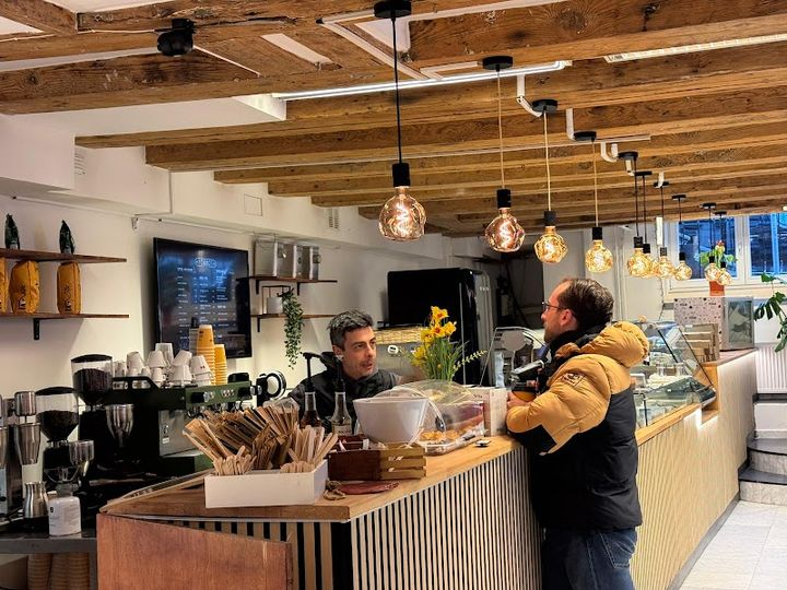 Inside Fournos: warm pendant lights, wooden counter, and a friendly barista serving a guest